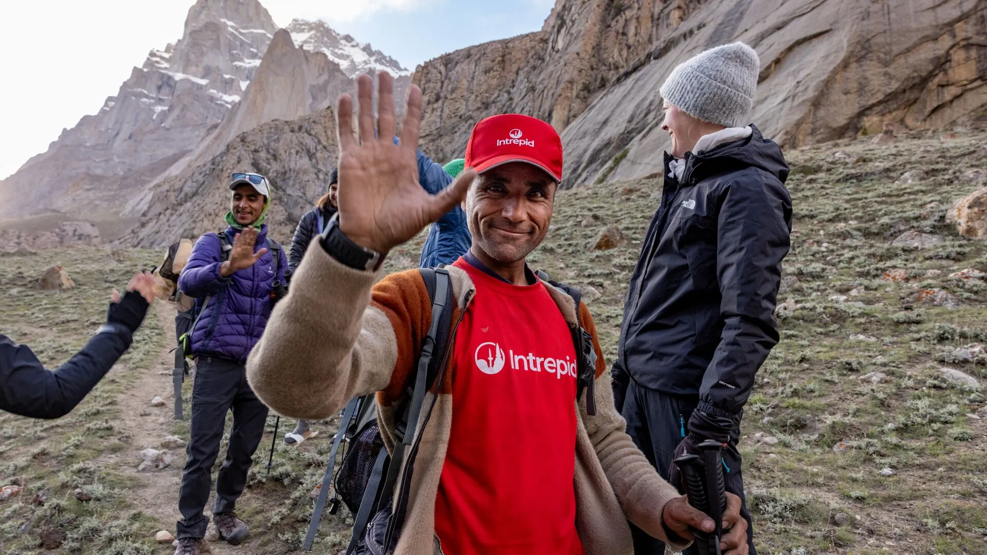 A smiling guide in a red Intrepid shirt waves to hikers in a mountainous landscape, promoting adventure travel.